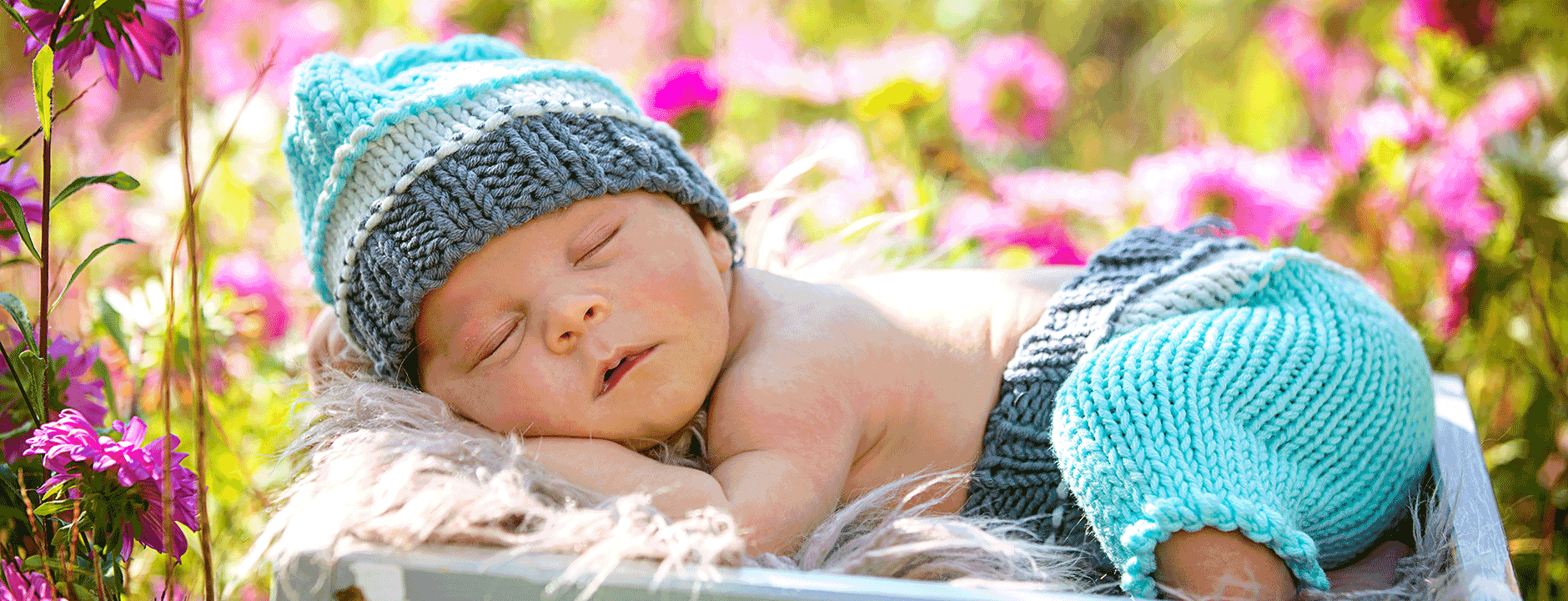 Schlafendes Baby in blau gestrickter Mütze und Hose in Geschenkbox mit Blumen zur Geburt